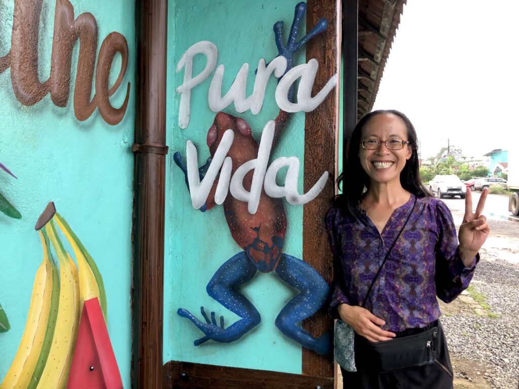 A photo of Sophy standing next to a mural that says "Pura Vida" and has a painting of a red jeans rainforest frog.